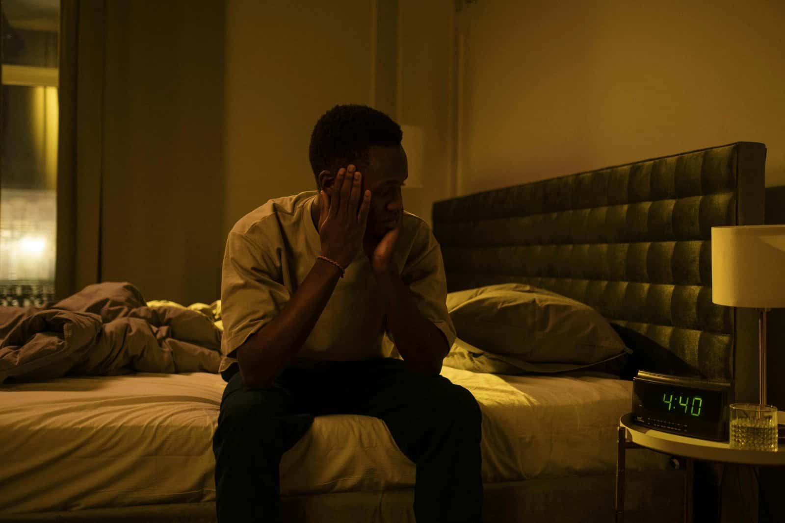 A man sitting on the edge of his bed at night with his head in his hands, a clock glowing on the nightstand