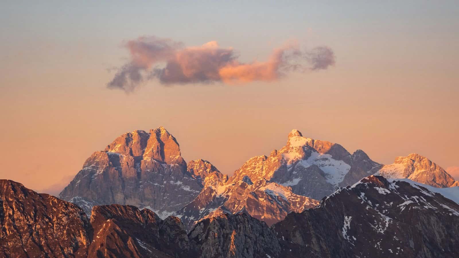 Rocky mountain peaks glowing orange and pink at sunset under a single small cloud