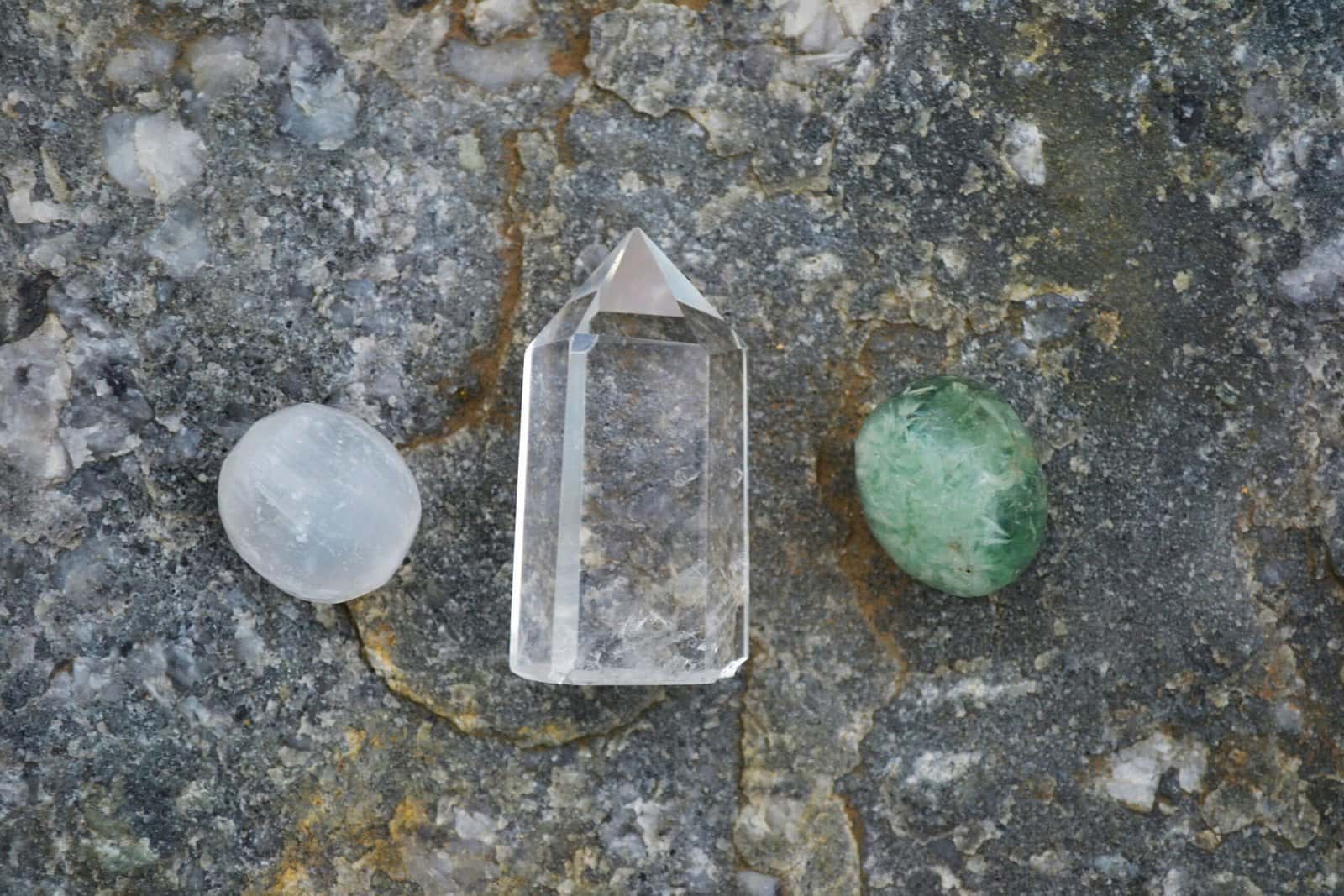 Three crystals, a clear quartz point between a white stone and a green stone, resting on grey rock