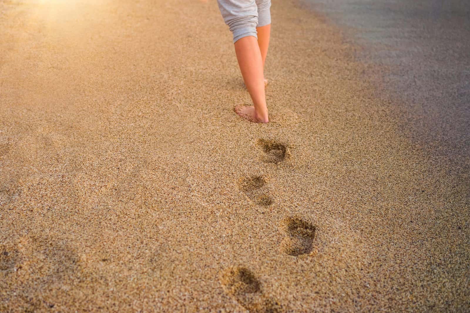 Bare feet walking along a sandy beach at sunset, leaving footprints behind